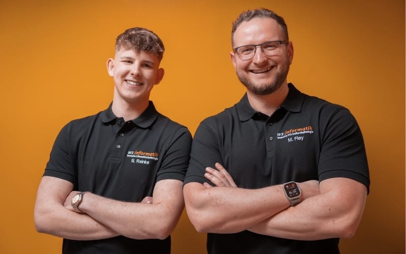 „Benedikt (links), ausgelernter Fachinformatiker für Systemintegration, und Michael (rechts), Servicetechniker bei der WS-Informatik GmbH & Co. KG, stehen Seite an Seite vor orangefarbenem Hintergrund – beide mit verschränkten Armen, im schwarzen Firmen-Poloshirt und mit einem Lächeln. Das Bild vermittelt Teamgeist, Kompetenz und Offenheit für neue Talente.“