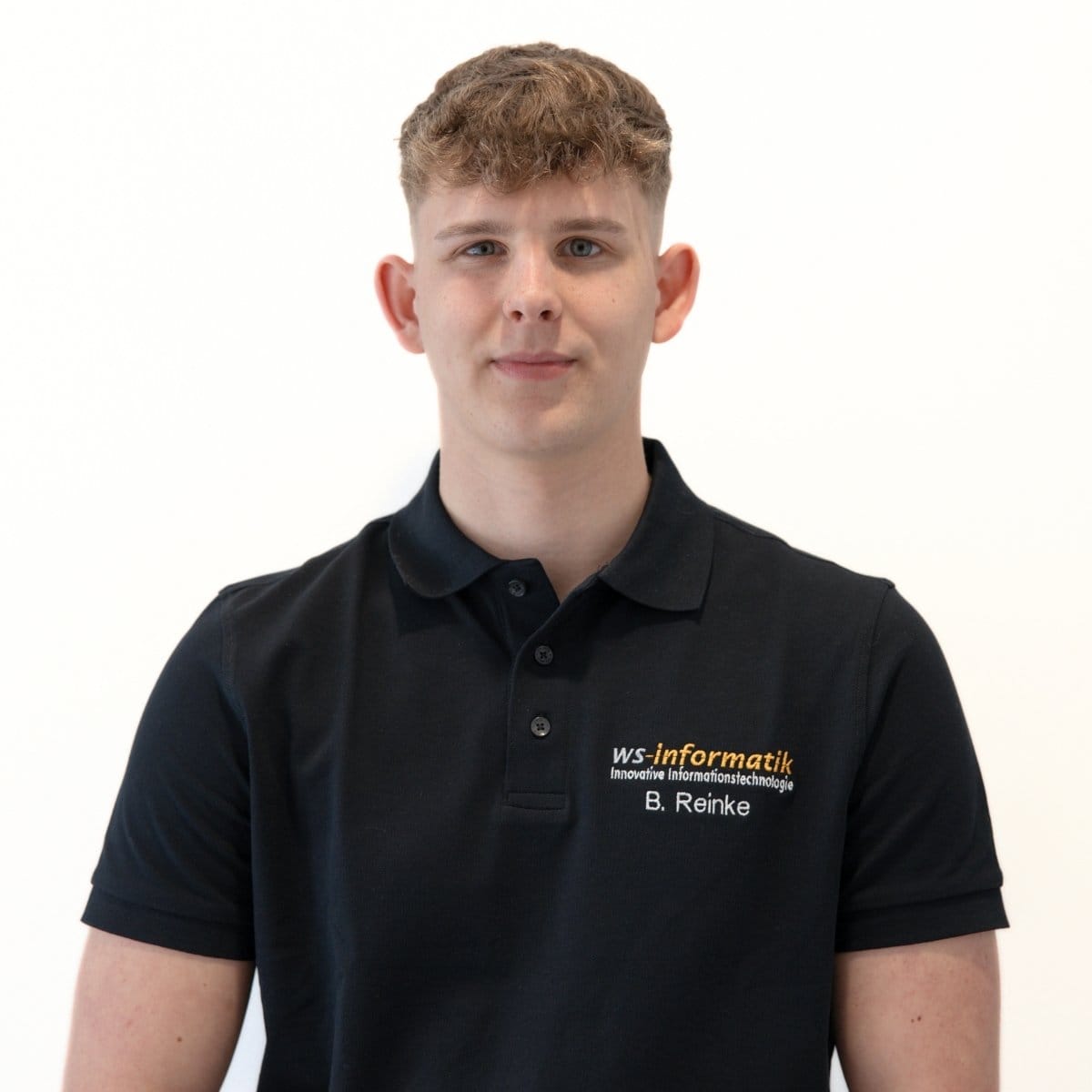 Portrait von Benedikt, Auszubildender als Fachinformatiker für Systemintegration bei der WS-Informatik GmbH & Co. KG, im schwarzen Poloshirt mit Firmenlogo, vor weißem Hintergrund.