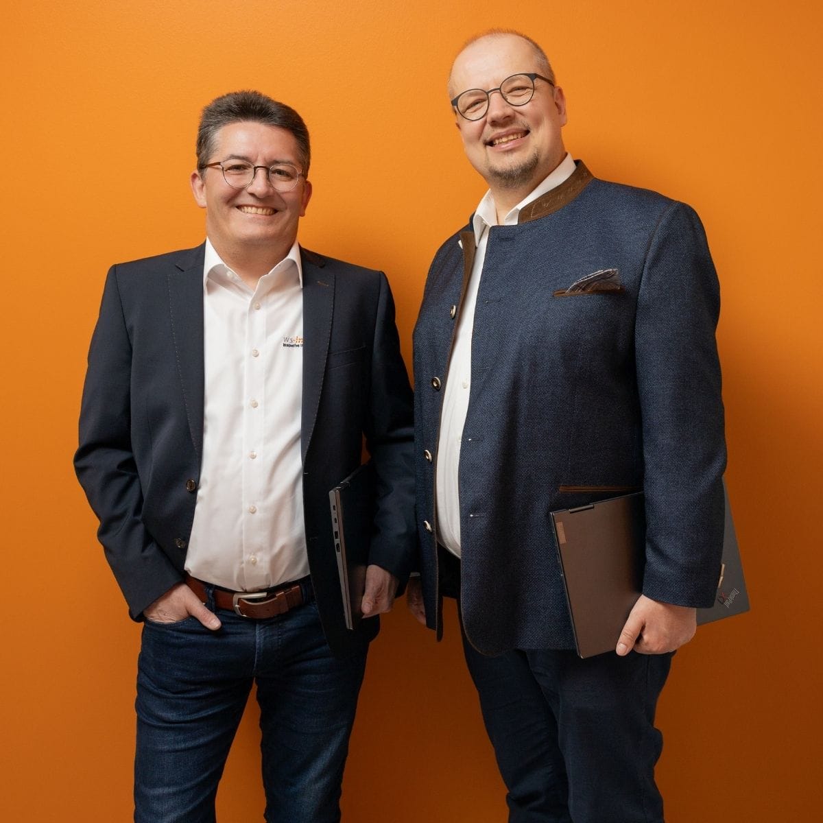 Dr. Stephan Werner (links) und Jan Steinhadt (rechts), Geschäftsführer der WS-Informatik GmbH & Co. KG, Seite an Seite vor orangefarbener Wand, beide mit Laptop in der Hand – ein starkes Team mit klarer Vision und gemeinsamem Antrieb.