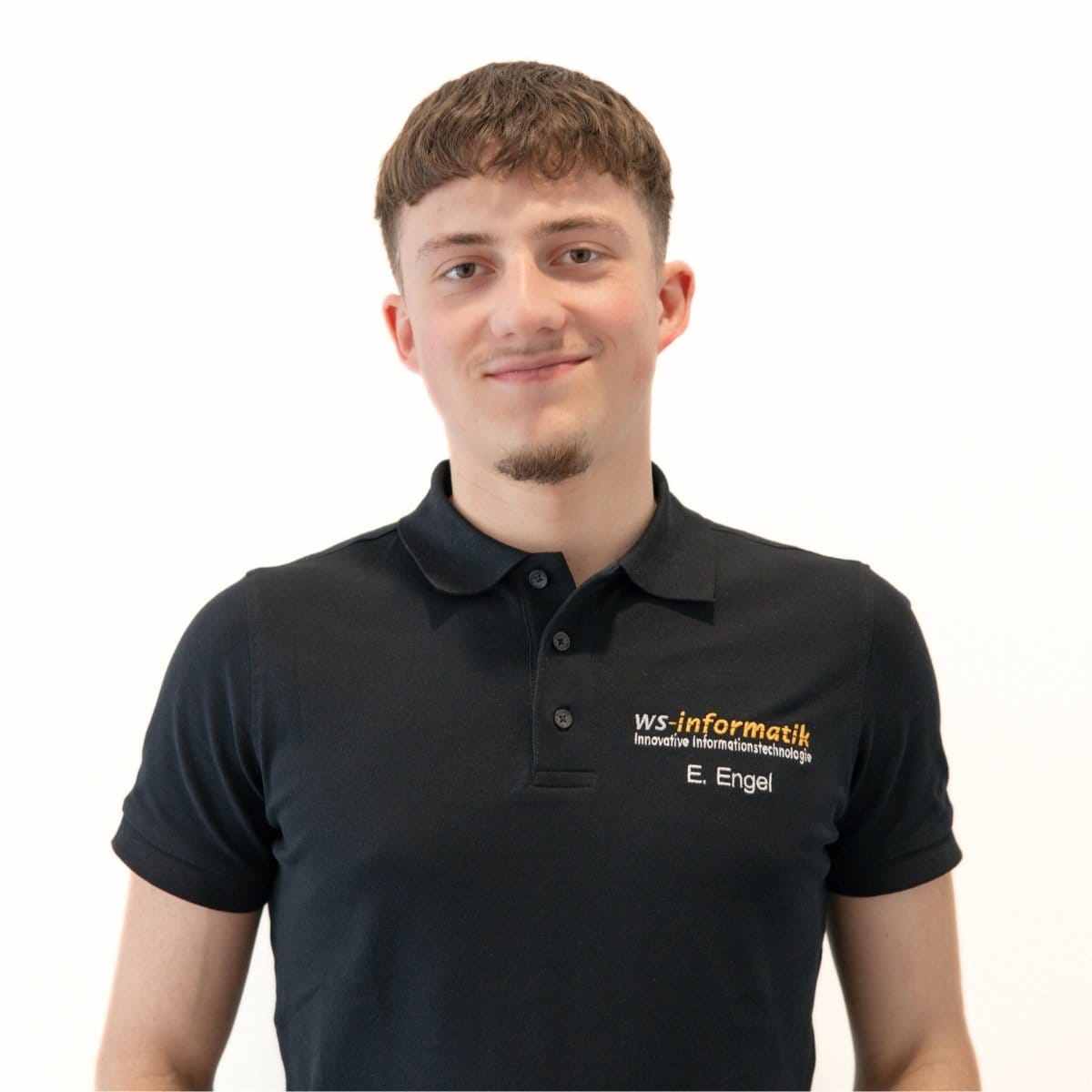 Portrait von Elias, Servicetechniker bei der WS-Informatik GmbH & Co. KG, im schwarzen Poloshirt mit Firmenlogo, vor weißem Hintergrund.