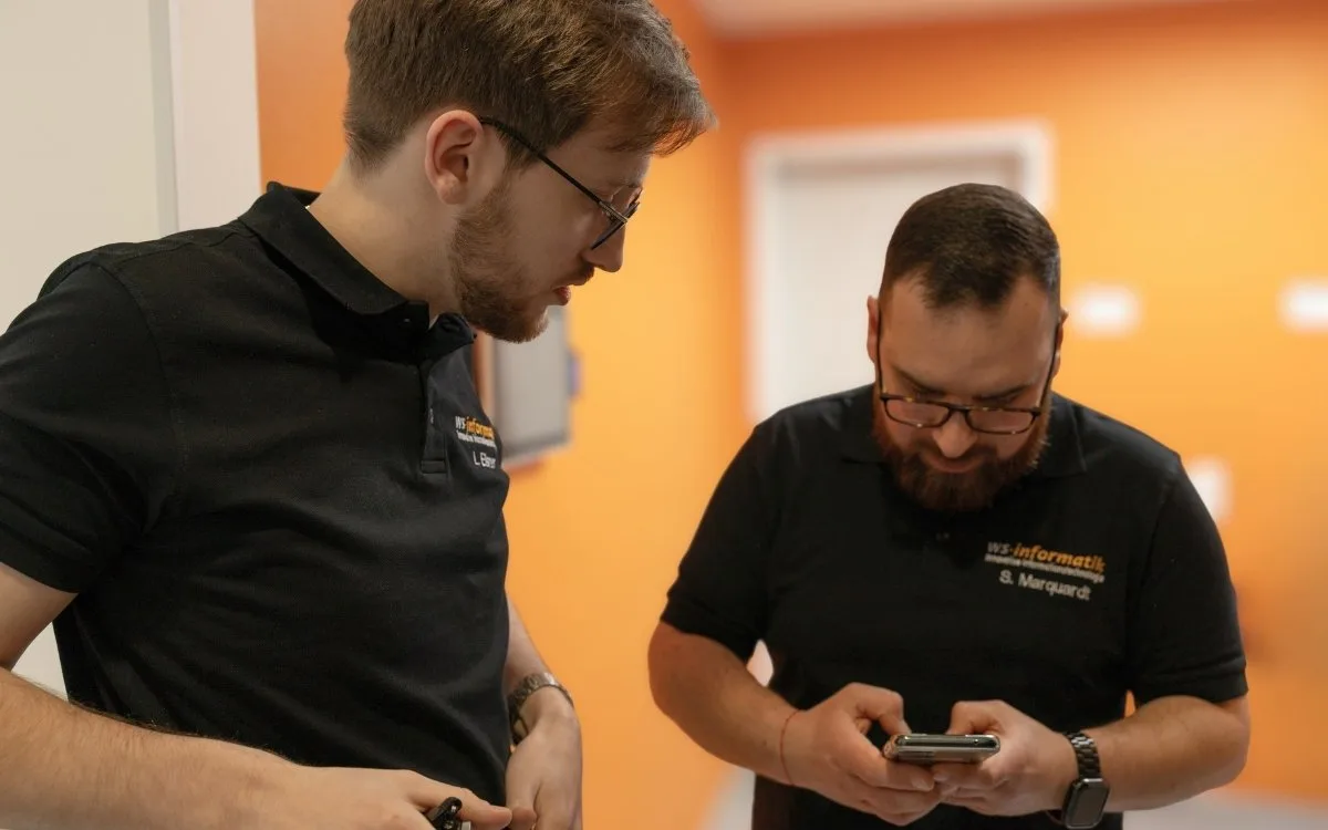Zwei Mitarbeitende der WS-Informatik im fachlichen Austausch auf einem Büroflur, einer zeigt eine Information auf dem Smartphone, der andere hört aufmerksam zu. Das Bild steht symbolisch für die professionelle Beratung, Beschaffung und Einrichtung von Hardware, Software und Arbeitsgeräten im Rahmen einer sicheren und durchdachten IT-Infrastruktur.