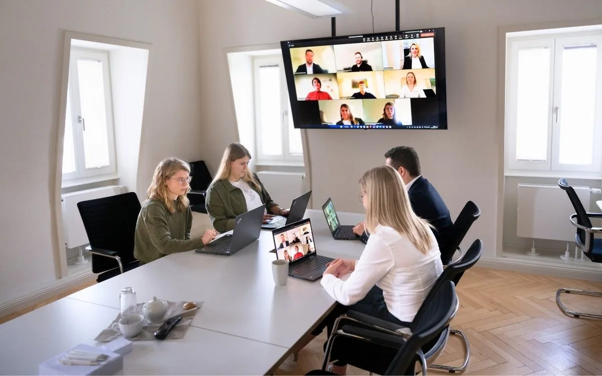 Das Bild zeigt eine Besprechungssituation in einem hellen, modernen Meetingraum. Mehrere Personen – sowohl Männer als auch Frauen – sitzen an einem langen Konferenztisch. Vor ihnen stehen geöffnete Laptops, auf den Bildschirmen ist eine Microsoft Teams-Besprechung mit mehreren zugeschalteten Teilnehmenden zu sehen. Die Personen im Raum wirken konzentriert und sind in die Videokonferenz eingebunden. Das Bild vermittelt professionelle digitale Zusammenarbeit, wie sie mit Lösungen von WS-Informatik möglich ist – etwa durch IP-basierte Telefonanlagen und nahtlos integrierte Microsoft Teams-Kommunikation.