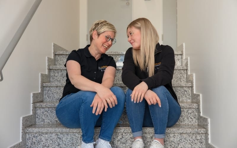 Sandra (links), Mitarbeiterin, und Lea (rechts), Assistenz der Geschäftsführung bei der WS-Informatik GmbH & Co. KG, sitzen lächelnd nebeneinander auf einer Treppe im Firmengebäude. Beide tragen schwarze Firmenpoloshirts und Jeans. Das Bild strahlt Kollegialität, Vertrauen und ein freundliches Arbeitsklima aus.