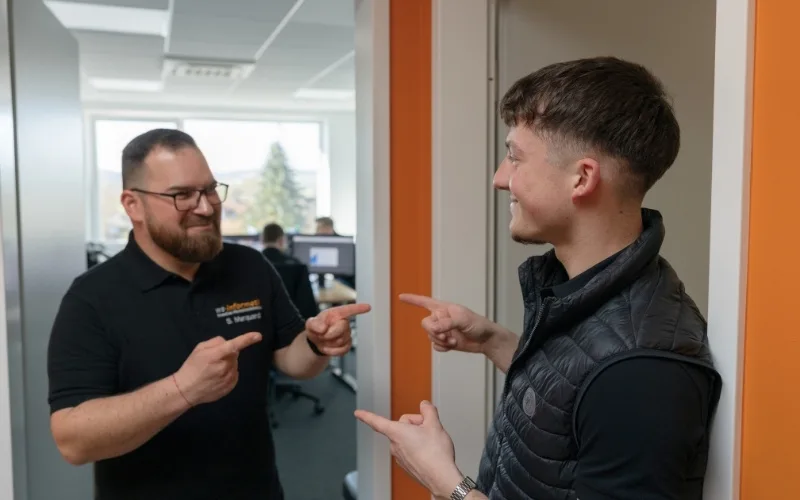 Sven (links), Servicetechniker, und Elias (rechts), Auszubildender als Fachinformatiker für Systemintegration bei der WS-Informatik GmbH & Co. KG, stehen sich in einem Büroflur gegenüber und zeigen lachend aufeinander. Das Bild vermittelt Kollegialität, Freude an der Arbeit und ein positives Miteinander im Team.