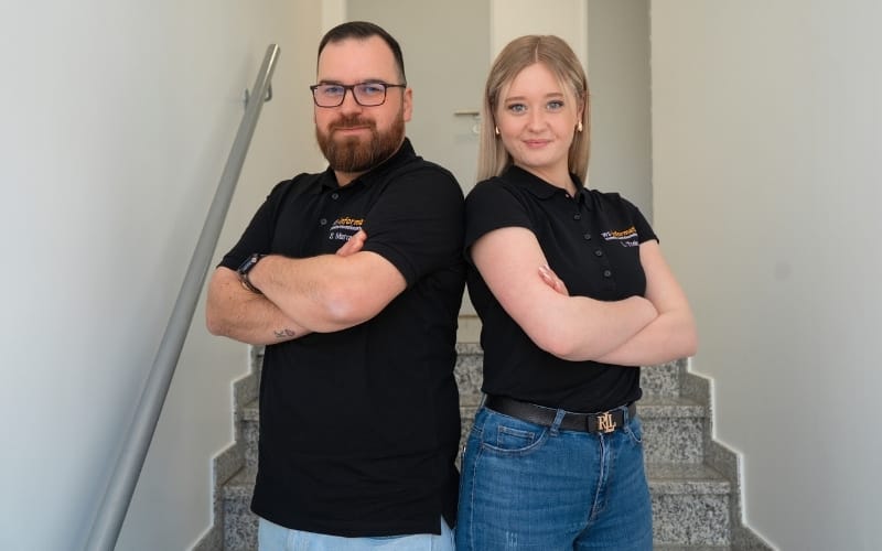 Sven (links), Servicetechniker, und Lea (rechts), Assistenz der Geschäftsführung bei der WS-Informatik GmbH & Co. KG, stehen Rücken an Rücken mit verschränkten Armen auf einer Treppe. Beide tragen schwarze Poloshirts mit Firmenlogo und blicken selbstbewusst in die Kamera – ein Bild für Teamgeist, Kompetenz und Engagement.